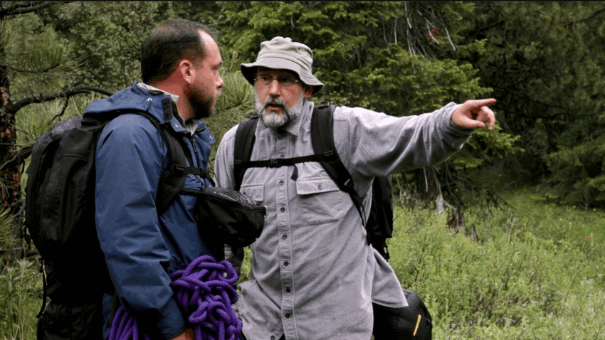 "Dalton, a rugged outdoorsman in a flannel shirt, and Kline, a skeptical academic in a blazer, heatedly argue about the logistics of landing a helicopter in a nearby forest clearing. The tension is palpable as Dalton gestures emphatically while Kline points to a map spread across a tree stump." The Ultimate Cult Bigfoot Adventure Movie